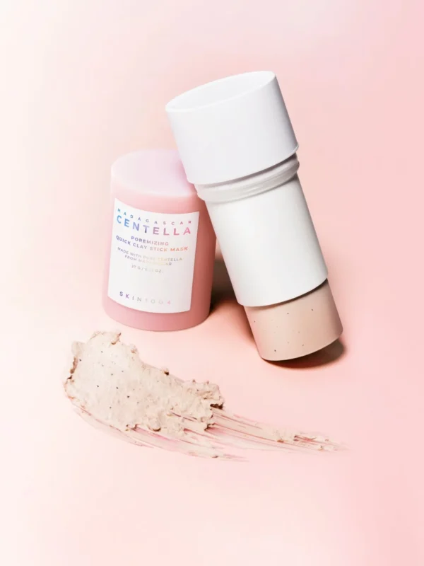Deux sticks du Madagascar Centella Poremizing Quick Clay Stick Mask, l’un ouvert montrant la texture beige rosée du masque, accompagnés d’une couche étalée d’argile au premier plan.