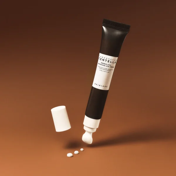Crème pour les yeux Madagascar Centella Probio-cica Bakuchiol Eye Cream de Skin1004 flottant sur fond brun avec bouchon détaché et gouttes de crème disposées au sol, mettant en avant l’embout applicateur précis.